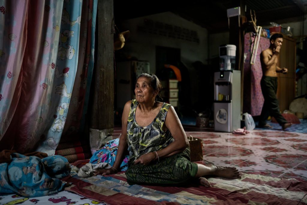 B&agrave; Amkha Janlong xem TV trong l&uacute;c con rể, Wittaya Thongnet, đi ph&iacute;a sau, trong ng&ocirc;i nh&agrave; họ sống b&ecirc;n s&ocirc;ng Mekong. Ảnh: The New York Times.
